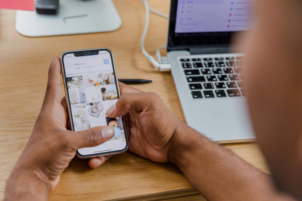Close-up of a person working on a smartphone while using a laptop at a wooden desk.